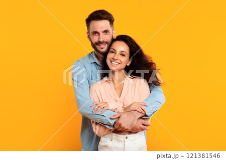 A man with a beard is embracing a woman with long dark hair. They are both smiling and looking directly at the camera. They are standing in front of a solid yellow background. A man with a beard is embracing a woman with long dark hair. They are both smiling and looking directly at the camera. They are standing in front of a solid yellow background. 121381546