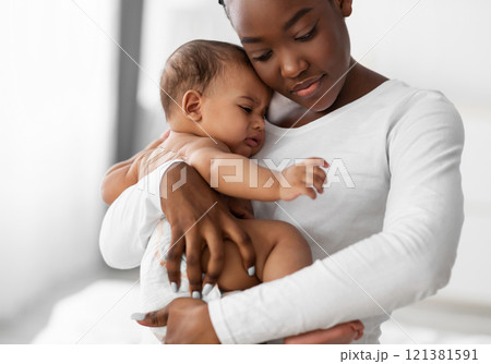 Maternity Concept. Portrait of young African American mother hugging and holding her upset crying baby on hands. Displeased hungry infant whining, feeling pain or colic. Copy space, blurred background Maternity Concept. Portrait of young African American mother hugging and holding her upset crying baby on hands. Displeased hungry infant whining, feeling pain or colic. Copy space, blurred background 121381591