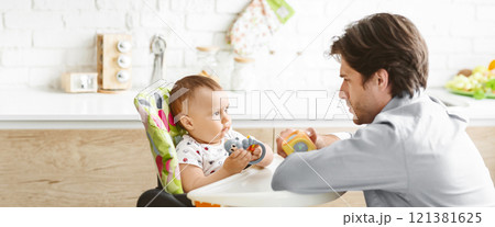 A father and his baby are playing together in the kitchen. The baby is sitting in a high chair with a colorful, patterned seat and is holding a toy. The father is sitting opposite the baby A father and his baby are playing together in the kitchen. The baby is sitting in a high chair with a colorful, patterned seat and is holding a toy. The father is sitting opposite the baby 121381625