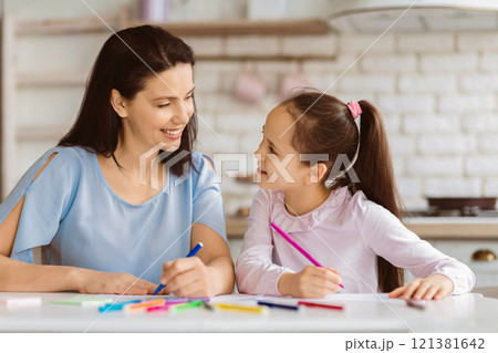 A mother and her young daughter sit at a table in their kitchen, coloring with colored pencils. They are looking at each other and smiling, enjoying a moment of bonding and creativity. 121381642
