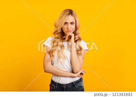 A young blonde woman in a white t-shirt and blue jeans stands in front of a bright yellow background. She has her hand on her chin and looks directly at the camera with a thoughtful expression. A young blonde woman in a white t-shirt and blue jeans stands in front of a bright yellow background. She has her hand on her chin and looks directly at the camera with a thoughtful expression. 121381681