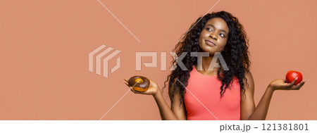 African American young woman with long curly hair, wearing a pink tank top, standing in front of a peach-colored background. She is holding a glazed donut in one hand and a red apple in the other African American young woman with long curly hair, wearing a pink tank top, standing in front of a peach-colored background. She is holding a glazed donut in one hand and a red apple in the other 121381801