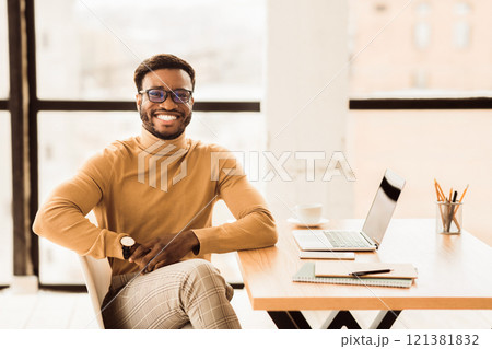 Enjoying Work. Smiling afro guy working on his laptop and looking at camera sitting at his working place, copyspace Enjoying Work. Smiling afro guy working on his laptop and looking at camera sitting at his working place, copyspace 121381832