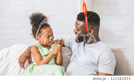 African American young girl wearing a crown and holding a makeup brush. She is sitting on the couch with her dad, who is wearing a red and orange headband. Both of them are smiling 121381833