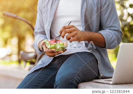 Unrecognizable African Man Eating Salad, Having Healthy Lunch Outdoors On Bench In Park, Cropped Image, Closeup, Selective Focus On Food Unrecognizable African Man Eating Salad, Having Healthy Lunch Outdoors On Bench In Park, Cropped Image, Closeup, Selective Focus On Food 121381844