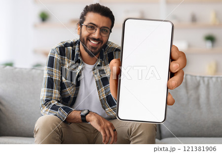 Cheerful Indian Guy Showing Smartphone With Big Blank White Screen At Camera, Happy Young Eastern Man Recommending New Mobile App Or Website While Sitting On Couch At Home, Creative Collage, Mockup 121381906