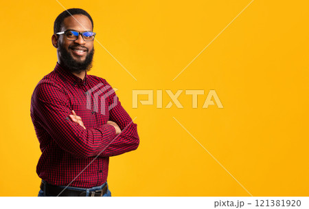 Confident Person. Portrait of smiling african american man wearing protective blue light glasses for computer, standing with folded arms isolated over yellow studio background wall with copy space 121381920
