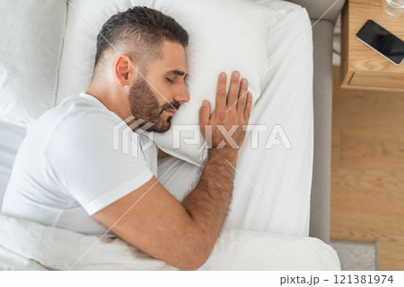 Top view of bearded man sleeping alone in white bed at home, his arm is placed next to his face. Resting concept 121381974