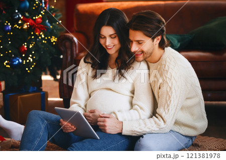 Pregnant cheerful woman showing her husband online kids store on digital tablet, sitting by xmas tree at home, shopping online, copy space 121381978