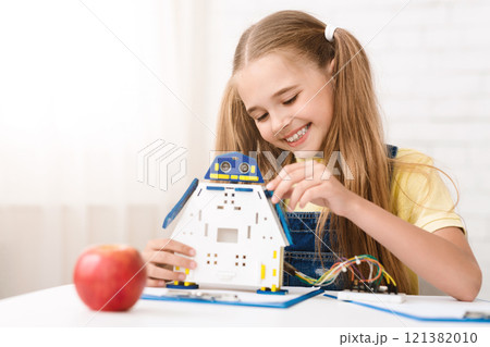 A young girl with pigtails smiles as she carefully assembles a robot toy at a table. She is wearing a yellow shirt and denim overalls. The robot is white with blue and yellow accents. A young girl with pigtails smiles as she carefully assembles a robot toy at a table. She is wearing a yellow shirt and denim overalls. The robot is white with blue and yellow accents. 121382010