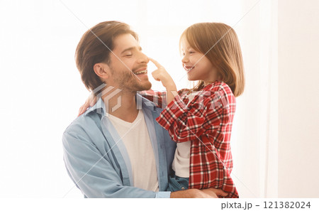 Portrait of cheerful father and little daughter bonding over white background at home, girl touching daddy nose 121382024
