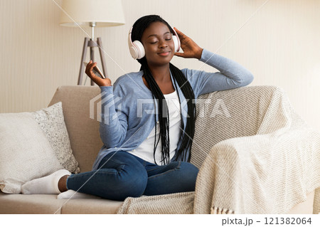 Favourite Leisure. Relaxed Black Girl Wearing Wireless Headphones, Listening Music On Smartphone, Sitting On Couch At Home Favourite Leisure. Relaxed Black Girl Wearing Wireless Headphones, Listening Music On Smartphone, Sitting On Couch At Home 121382064