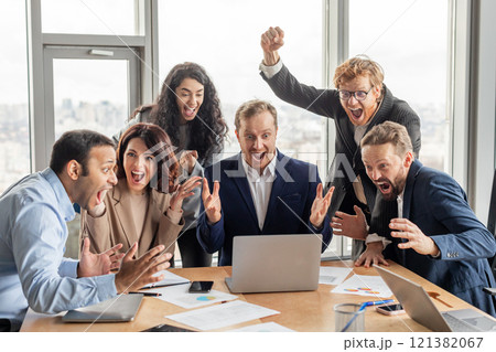 A diverse group of business professionals celebrates a successful project or achievement. They are gathered around a laptop, all wearing professional attire and beaming with joy 121382067