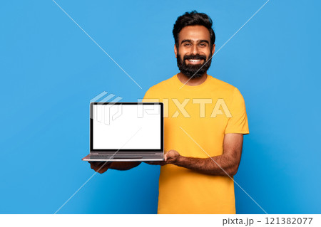 A bearded Indian man in a yellow t-shirt smiles while holding a laptop with a blank screen in front of him. He is standing in front of a solid blue background. A bearded Indian man in a yellow t-shirt smiles while holding a laptop with a blank screen in front of him. He is standing in front of a solid blue background. 121382077