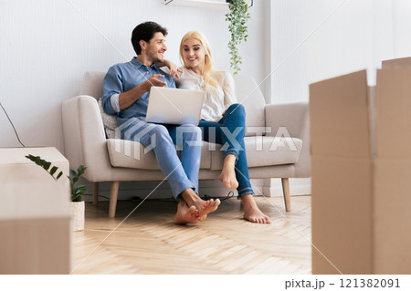 A young couple sits on a couch in their new apartment, smiling and looking at a laptop. 121382091