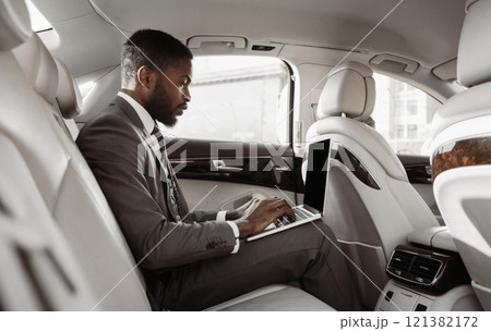African American businessman sits in the backseat of a car, working on his laptop computer. He is wearing a suit and tie and appears to be focused on his work African American businessman sits in the backseat of a car, working on his laptop computer. He is wearing a suit and tie and appears to be focused on his work 121382172