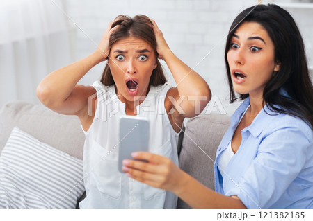 Omg. Shocked Girls Looking At Smartphone Screen Reading Message Sitting On Couch Indoor. Selective Focus 121382185