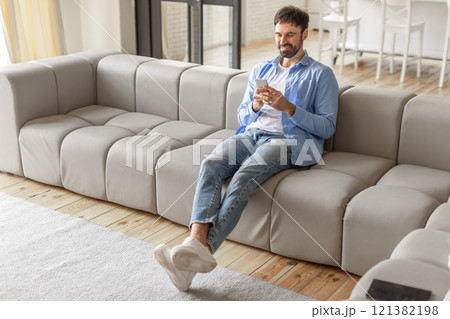 A man sits on a light grey modular couch in a living room, relaxed and smiling as he uses his phone. The man is dressed casually in a blue button-down shirt, white t-shirt, jeans, and white sneakers 121382198