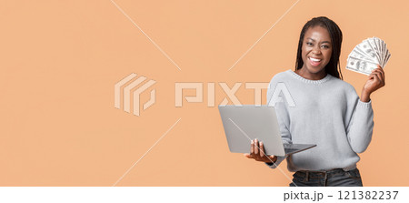 E-commerce and profit online. Young successful black woman making money from internet, holding cash and laptop in hands, panorama with copy space, beige background 121382237