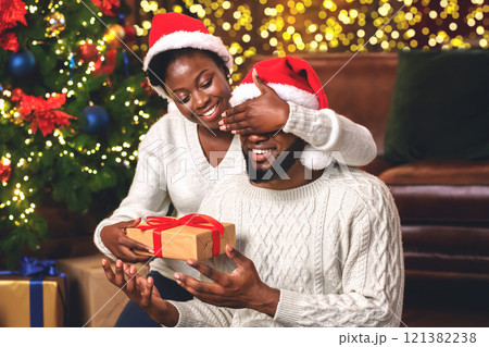 Surprise for Christmas. Smiling african american woman in Santa hat closing eyes to her boyfriend while giving him present near Xmas tree 121382238