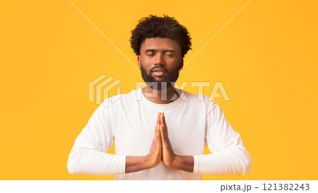Concentrated black man with closed eyes holding his hands in praying gesture, orange background Concentrated black man with closed eyes holding his hands in praying gesture, orange background 121382243
