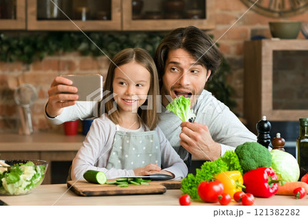 Cooking Selfie. Joyful Young Dad Taking Photo On Smartphone With His Little Daughter In Kitchen While They Preparing Healthy Lunch Together, Panorama Cooking Selfie. Joyful Young Dad Taking Photo On Smartphone With His Little Daughter In Kitchen While They Preparing Healthy Lunch Together, Panorama 121382282