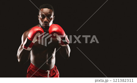 A muscular male boxer wearing red boxing gloves stands with a determined expression, ready for a fight in a dark studio. A muscular male boxer wearing red boxing gloves stands with a determined expression, ready for a fight in a dark studio. 121382311