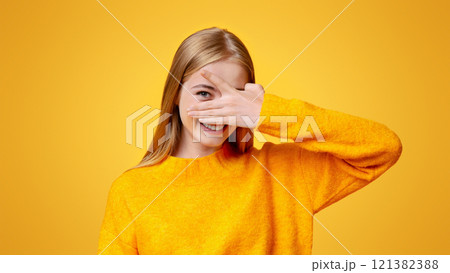 Playful teen girl covering face with hand and peeking through fingers over orange background with copy space 121382388