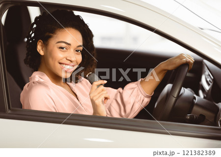 Joyful Afro Woman Showing Car Key Smiling To Camera Sitting In Driver's Seat In New Automobile Dealership Store. Selective Focus 121382389