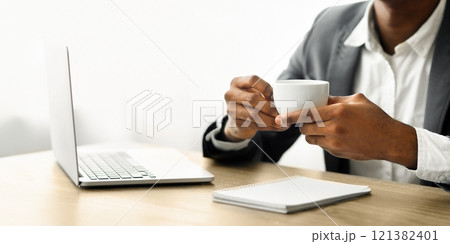 A businessman sits at a desk with a laptop and notebook, holding a white coffee cup. 121382401