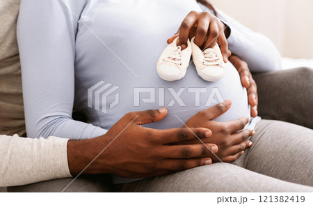 Close up of pregnant black woman belly with baby shoes on it, husband hugging his wife, new family concept 121382419