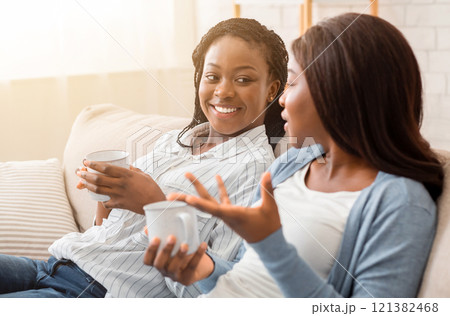 Two female friends sitting together on sofa chatting and drinking tea, having fun at home. Two female friends sitting together on sofa chatting and drinking tea, having fun at home. 121382468