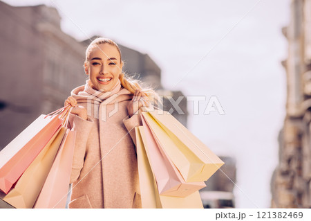 A young woman is walking on a city street with shopping bags in her hands. She is smiling and looks happy. The background is blurred, and the weather is sunny, copy space 121382469