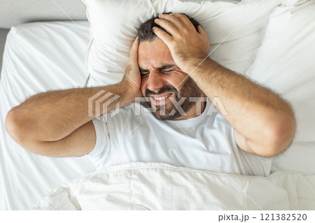 A man lies in bed, his face contorted in pain as he holds his head with both hands. He is likely suffering from a headache or migraine, his expression showing discomfort and distress, top view 121382520