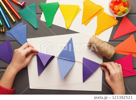 Birthday handmade background. Female hands making party paper decorations and colorful flags garland. DIY accessories on gray wooden planks, top view. Holiday organization, hobb, handicraft concept 121382521