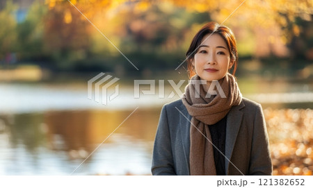 A woman with dark hair wearing a brown scarf and grey coat stands in a park. The background is blurry with a pond and yellow, orange, and red leaves. 121382652