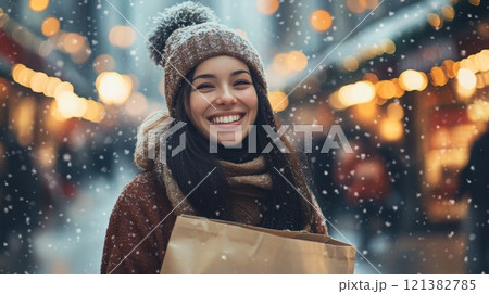Snowflakes gently fall as a woman enjoys her time in a lively winter market, wearing a warm sweater and beanie. She holds shopping bags and beams with happiness amidst twinkling lights. 121382785