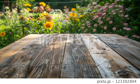 A wooden table sits in front of a garden filled with blooming flowers in shades of yellow, pink, and white. The table is weathered and shows signs of age, with a rustic charm. A wooden table sits in front of a garden filled with blooming flowers in shades of yellow, pink, and white. The table is weathered and shows signs of age, with a rustic charm. 121382804
