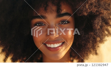 A young woman with vibrant, curly hair smiles brightly while standing outdoors in warm sunlight, enjoying a moment of joy. 121382947