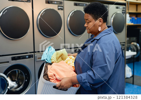 Side view of senior African American woman carrying basket of laundry preparing colored clothes for washing at self service laundromat, copy space Side view of senior African American woman carrying basket of laundry preparing colored clothes for washing at self service laundromat, copy space 121384328