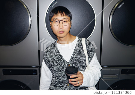 Portrait of modern Asian man with reusable cup in hand standing against washing machines Portrait of modern Asian man with reusable cup in hand standing against washing machines 121384366