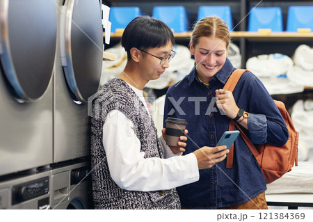 Medium shot of young Asian man showing video on phone to cheerful female friend while washing clothes and waiting for result at self service laundry 121384369
