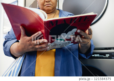 Cropped shot of senior African American woman reading fashion magazine while passing time in self service laundry room waiting for end of washing, copy space 121384375
