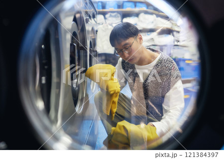 Shot through open washing machine of young Asian man unloading clean clothes while doing daily laundry at coin laundromat, household chores concept 121384397