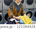 Cropped shot of young woman sorting colored sweaters on folding table preparing to load delicate clothes into washing machine in self service laundry, copy space 121384428