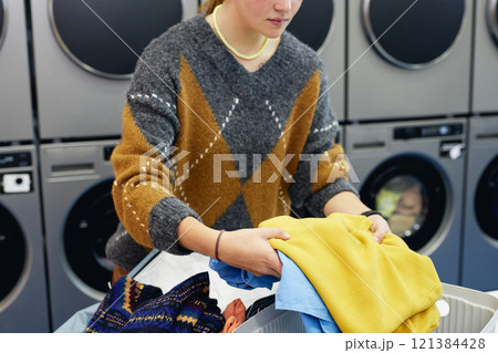 Cropped shot of young woman sorting colored sweaters on folding table preparing to load delicate clothes into washing machine in self service laundry, copy space 121384428