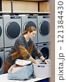 Vertical shot of young woman sorting through laundry in basket on folding table preparing to load clothes into washing machine in coin laundromat, copy space 121384430