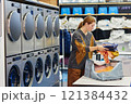 Side view of young woman putting pile of sweaters into basket on folding table while preparing to load clothes in washing machine in self service laundry room, copy space 121384432