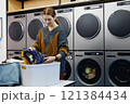 Medium shot of young woman holding vintage sweaters sorting through basket while checking result after washing in laundry machine in coin laundromat, copy space 121384434