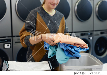 Cropped shot of young woman holding pile of colored clothes sorting delicate garment before loading into washing machine in self service laundry, copy space 121384436
